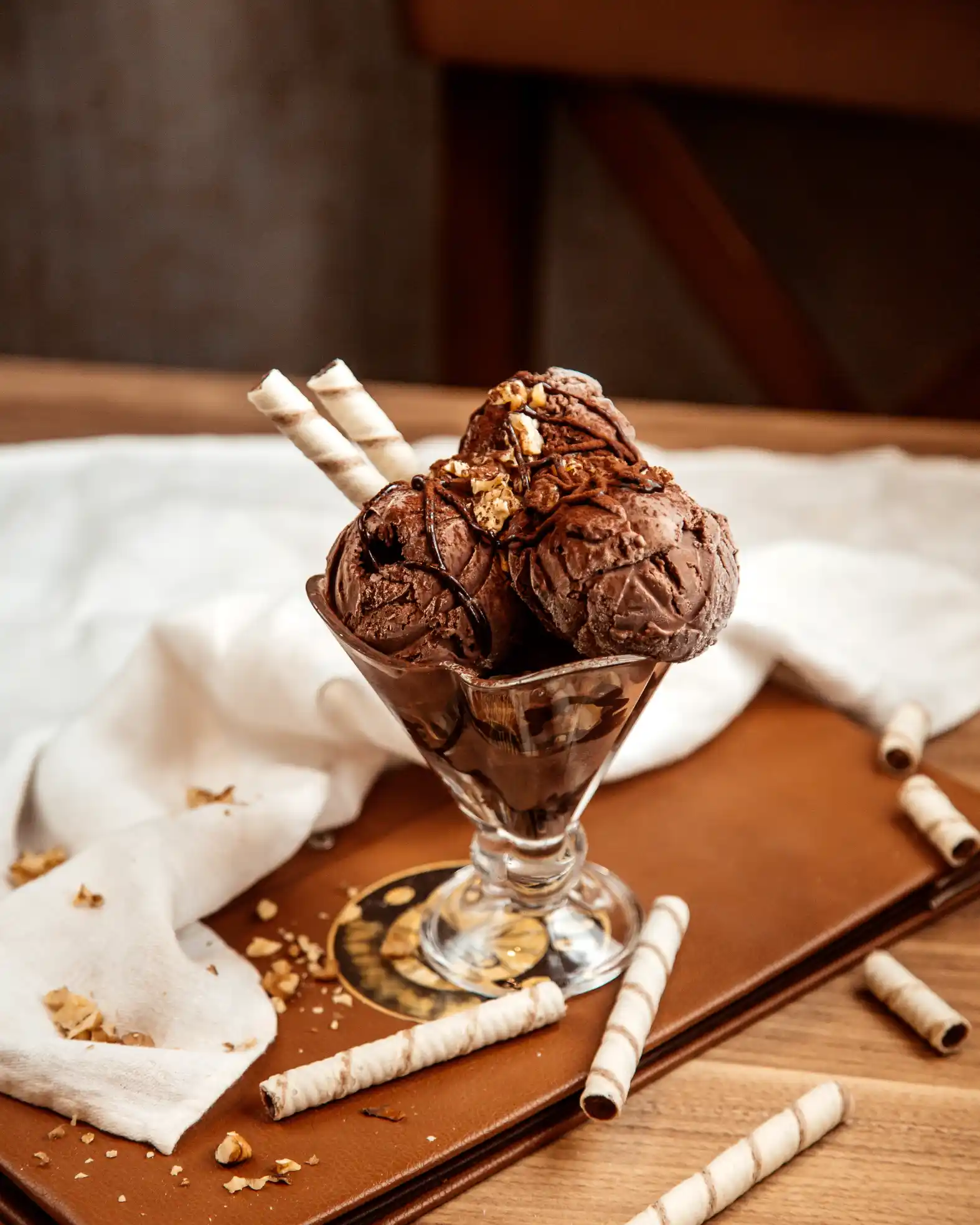 Como Vender Sorvete no Verão: taça com sorvete de chocolate cremoso, calda e pedaços ao lado de cones de massa e ingredientes sobre uma mesa de madeira, com ambiente aconchegante